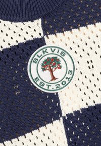 Gestrickter Pullover mit marineblauem und cremefarbenem Karomuster und offenem Mesh-Gewebe. Ein runder Aufnäher zeigt ein Baummotiv mit der Aufschrift "BLKVIS EST. 2013."