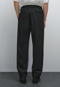Person wearing black tailored trousers and black shoes standing on dark speckled floor against light gray wall, viewed from behind.