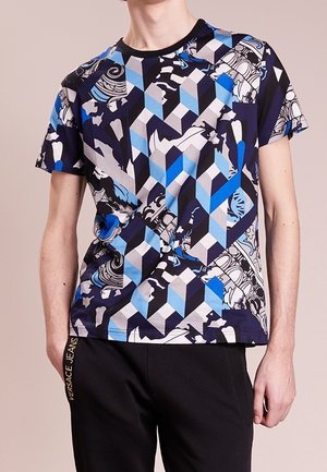 Male wearing a short-sleeve geometric patterned t-shirt in blue, black, and white with black pants featuring "Versace Jeans" in gold lettering.
