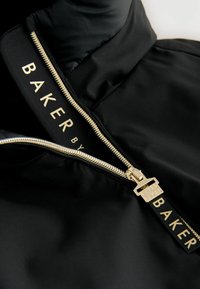 Črna jakna z visokim ovratnikom, opremljena z zlatim zadrgo in kontrastno oznako z logotipom z napisom "BAKER". Gladka, elegantna tekstura tkanine.
