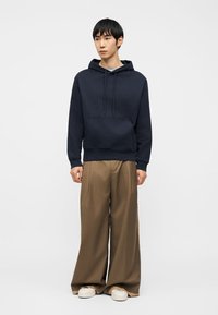 Άνδρας που φοράει μπλε-σκούρα φούτερ με κουκούλα, καφέ φαρδιά παντελόνια και άσπρα αθλητικά παπούτσια, στέκεται μπροστά σε απλό φωτεινό φόντο.