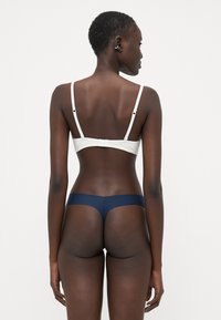 Witte bralette met dunne bandjes en een haaksluiting aan de achterkant, gecombineerd met een marineblauwe string. Glad materiaal, minimalistisch ontwerp, toont achterkant.