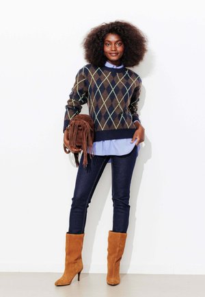 Femme aux cheveux naturels portant un pull à losanges, une chemise bleue, un jean foncé, des bottes en daim couleur sable, tenant un sac à main marron à franges, debout contre un mur blanc.