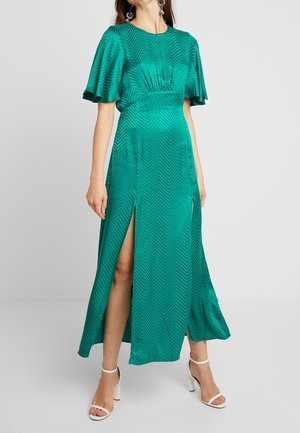 Vrouw draagt een satijnen jurk in teal met chevronpatroon, korte vlinderachtige mouwen en een hoge split aan de voorkant tot aan de dij, gecombineerd met witte sandalen met hak.