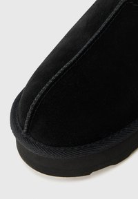 Zwarte suède slipper met een ronde neus, contrasterende zwarte stiksels en een textuurstof trim langs de rand van de zool.