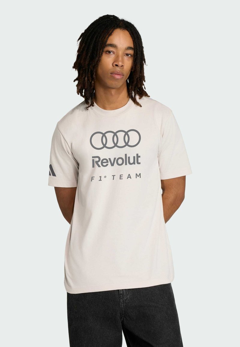 Beżowa bawełniana koszulka z logo Audi i napisem "Revolut F1 TEAM" w kolorze szarym. Krótkie rękawy, z subtelnymi czarnymi paskami na jednym rękawie.