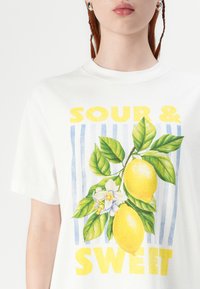 T-shirt in cotone bianco con una grafica di limone, foglie verdi e fiori bianchi, accompagnata dal testo "SOUR & SWEET" in giallo.