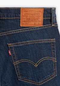 Dunkelblaue Jeans aus Denim mit einem braunen Lederetikett, das geprägten Text zeigt. Gelbe Nähte umrahmen das Design der Gesäßtasche.