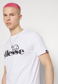 Hvit kortermet t-skjorte med et stort svart "ellesse"-logo på fronten, og et lite logo med fargeakenter på venstre erme.