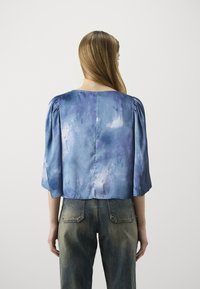 Blouse à manches courtes en soie bleu clair avec un motif tie-dye, dotée d'une coupe ample et de manches légèrement bouffantes. Portée avec un jean en denim délavé.
