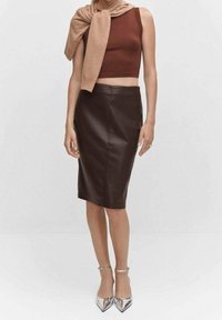 Jupe crayon en cuir marron associée à un crop top rouille et un pull beige noué autour des épaules. Des escarpins argentés à talons aiguilles complètent la tenue.