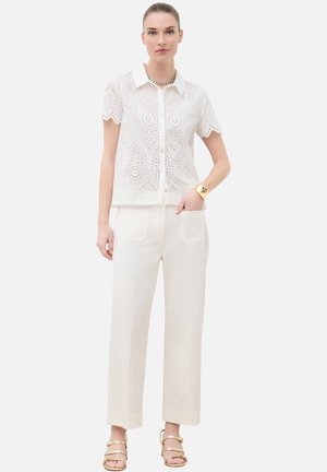 Femme portant un chemisier blanc à manches courtes en broderie anglaise boutonné, pantalon large crème, collier chaîne dorée, bracelet et sandales à lanières.