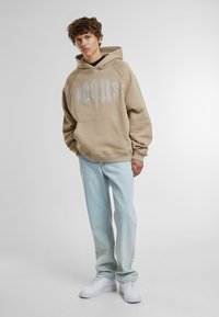 Felpa beige con tasca frontale e grande testo grigio. Abbinata a jeans azzurri chiari e sneakers bianche, presenta una vestibilità comoda.