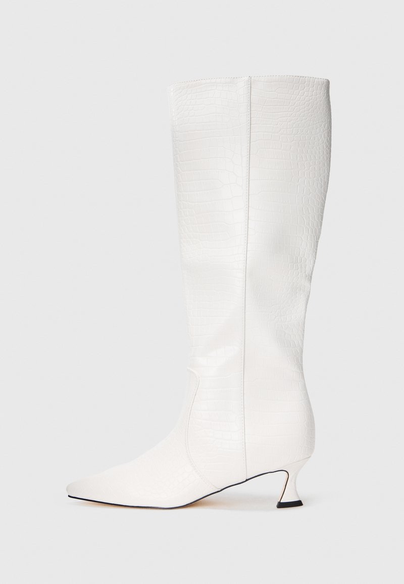 Bota blanca hasta la rodilla con textura de cocodrilo, punta afilada y tacón bajo curvado sobre un fondo blanco liso.