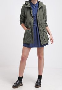 Olivově zelená parka s kapucí a zipem, se dvěma předními kapsami a volným střihem, navlečená přes modrou denimovou šaty a černé boty se sockami.