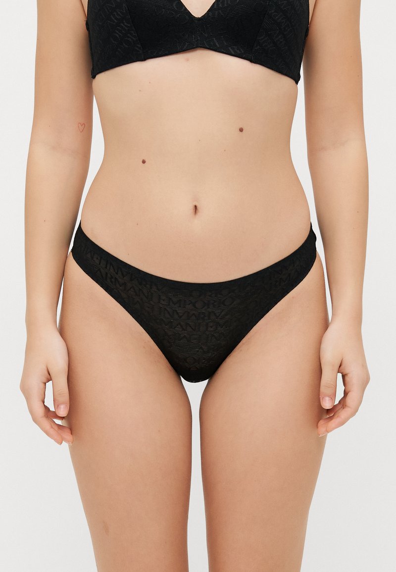 Conjunto de lencería negra que incluye un sujetador tipo bralette con escote en V y un tanga de corte bajo. Ambas piezas están confeccionadas con una tela texturizada que presenta un patrón repetido de logos.