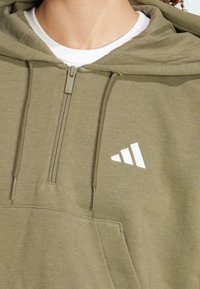 Olivgrön zip-up hoodie med en känguruficka, justerbara dragsko och en vit logotyp. Tillverkad av mjukt, texturerat tyg.
