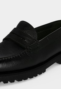 Mocassin noir en cuir texturé avec bride penny et semelle épaisse en caoutchouc à crampons, présenté en angle sur fond blanc.