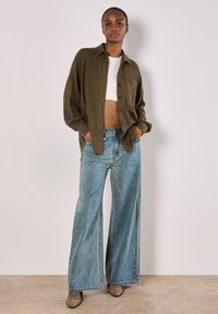 Olive green ribbed shirt, large fit, button-front, layered over a white crop top. Light blue wide-leg denim jeans, raw hem, paired with grey boots.