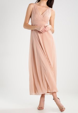 Vrouw die een mouwloze, geplooide maxi-jurk in blushroze draagt met een diepe V-hals en beige enkellaarsjes met bandje, staande tegen een effen achtergrond.