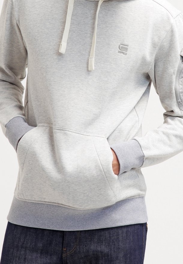 Personne portant un sweat à capuche gris clair avec poche avant et cordons crème, mains dans la poche, associé à un jean bleu foncé.