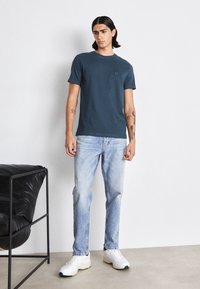 Granatowa koszulka z krótkim rękawem z kwadratową kieszenią na piersi, zestawiona z jasnoniebieskimi dżinsami i białymi sneakersami z zielonymi akcentami.