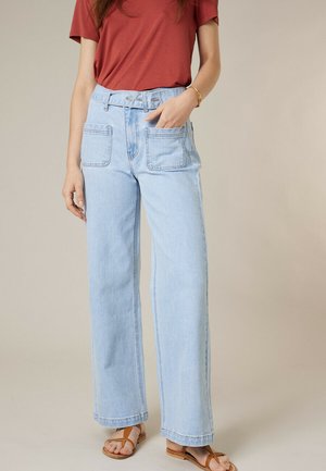 Femme portant un jean large bleu clair avec de grandes poches avant, un t-shirt couleur rouille et des sandales plates marron, posant devant un fond neutre.