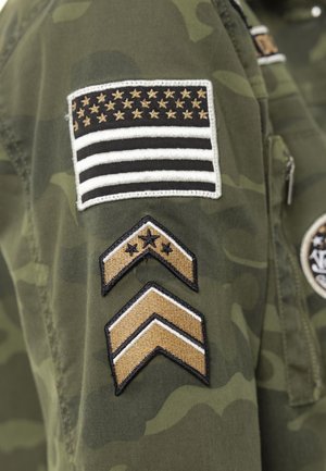 Manche de veste camouflage verte avec patch brodé du drapeau américain en noir et or, ainsi que deux insignes de grade en chevron en dessous.
