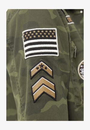 Manche de veste camouflage verte avec patch brodé du drapeau américain en noir et or, ainsi que deux insignes de grade en chevron en dessous.