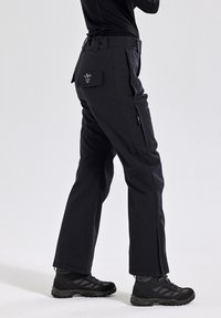 Zwarte buitenbroek met een rechte snit, ritszakken, een verstelbare taille en verstevigde naden. Gecombineerd met zwarte wandelschoenen.