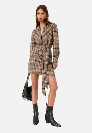 Nainen, jolla on yllään ruskea ruudullinen kietaisumekko, mustat nilkkurit, kädessään musta hapsullinen käsilaukku, seisoo yksiväristä taustaa vasten.