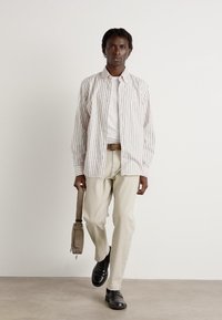 Camicia a righe bianca e beige chiaro con maniche lunghe, abbinata a pantaloni beige e scarpe nere, mentre tiene una borsa grigia.