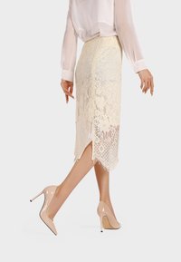 Gonna a tubino in pizzo crema con uno spacco alto, caratterizzata da motivi floreali e un orlo frangiato, abbinata a décolleté nude.