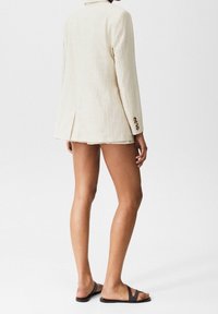 Blazer beige claro con rayas finas de diseño ajustado y una abertura frontal sola, combinado con pantalones cortos a medida y sandalias negras.
