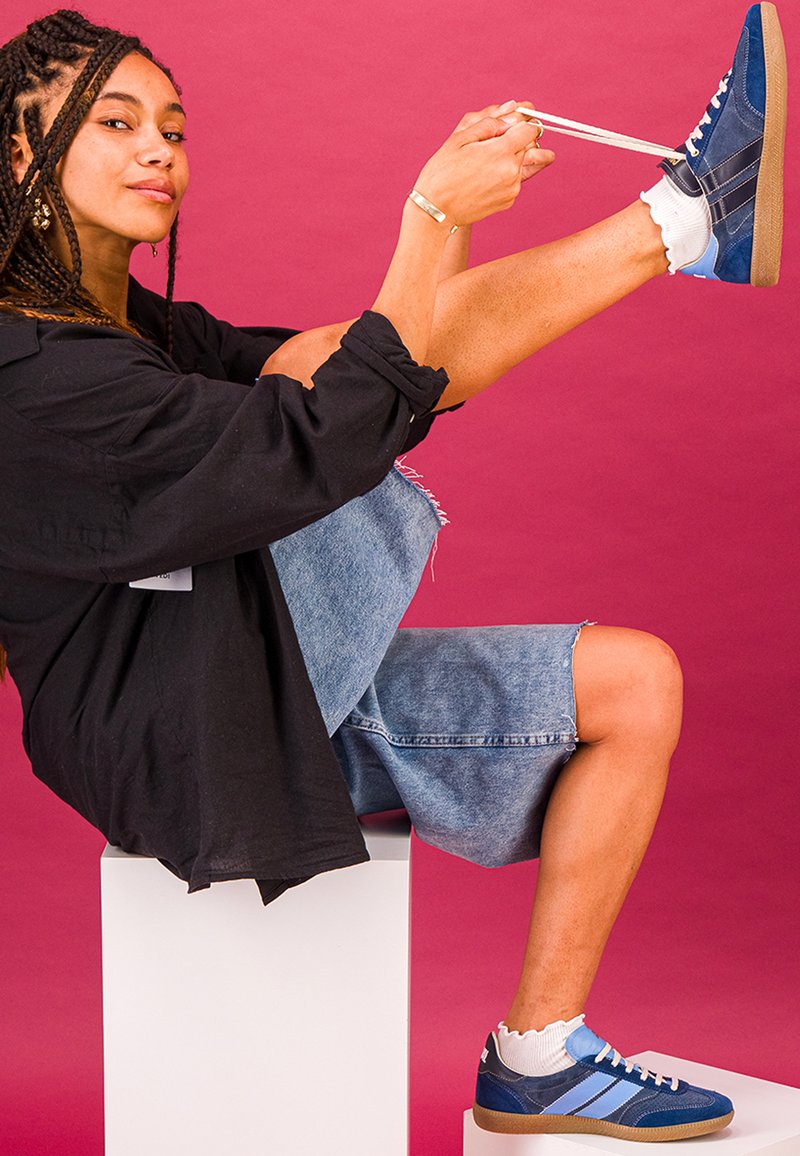 Femme aux cheveux tressés assise sur un bloc blanc, remontant la jambe pour nouer le lacet d'une basket bleue sur un fond rose.