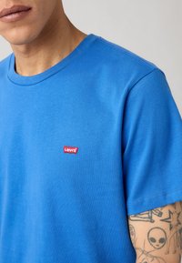 Κοντινό πλάνο ενός ατόμου που φοράει ένα φωτεινό μπλε μπλουζάκι Levi's με κόκκινο λογότυπο και ορατά τατουάζ στο χέρι, σε ουδέτερο φόντο.