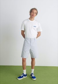 Lichtblauwe shorts met een tailored fit, een knoopsluiting en een vlakke voorkant. Draagt bij een wit t-shirt en blauwe sneakers. Effen witte achtergrond.