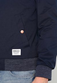 Marineblauwe jas met een opgezette zijzak met knopen, geribbelde grijze manchetten en een wit label met merknaam aan de zoom. Gladde stoftextuur.