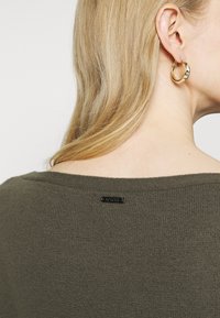 Oliwkowy sweter z prążkowanym dekoltem i czarną metką logo z tyłu; gładka faktura, dopasowany krój i krótkie rękawy.