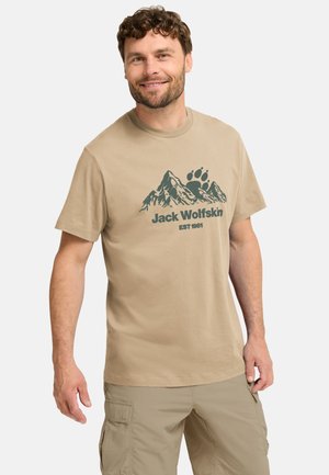 Beige katoenen T-shirt met een groene berglandschapgrafiek en de tekst "Jack Wolfskin EST 1981" op de voorkant gedrukt. Korte mouwen, ronde hals.