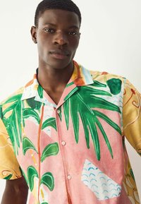 Camisa de manga corta con cuello que presenta un estampado tropical con hojas de palmera verdes y patrones abstractos en tonos amarillos, rosas y azules.