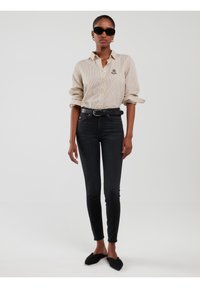 Femeie purtând ochelari de soare negri, cămașă cu dungi bej și nasturi, curea neagră, blugi negri cu talie înaltă și pantofi negri cu vârf ascuțit, pe fundal alb.