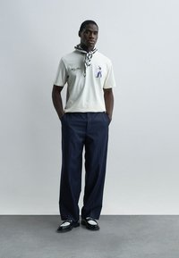 Άνδρας που φοράει λευκό μπλουζάκι με σχέδιο, ασπρόμαυρο κασκόλ, μπλε ναυτικό παντελόνι με ρίγες και ασπρόμαυρα μοκασίνια, στέκεται μπροστά σε απλό φόντο.