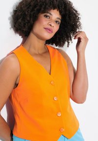 Oranje mouwloze vest met een v-halslijn, vijf oranje knopen aan de voorkant, gecombineerd met lichtblauwe broek. Glad materiaal en gestructureerd ontwerp.