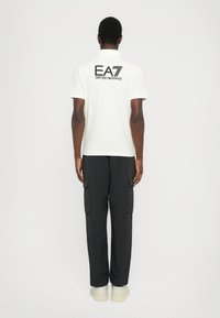 Bela polo majica z logotipom EA7 Emporio Armani na hrbtni strani, v kombinaciji z črnimi cargo hlačami in svetlimi športnimi copati. Gladek material in sproščen kroj.