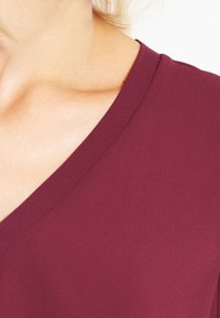 Haut bordeaux à col en V avec une texture lisse et douce, montrant l'encolure et l'épaule. Le tissu semble léger avec un léger éclat.