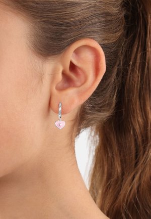 Orecchini a cerchio in argento con un piccolo charm a forma di cuore rosa con un gemma centrale, appesi a un lobo dell'orecchio su uno sfondo neutro.