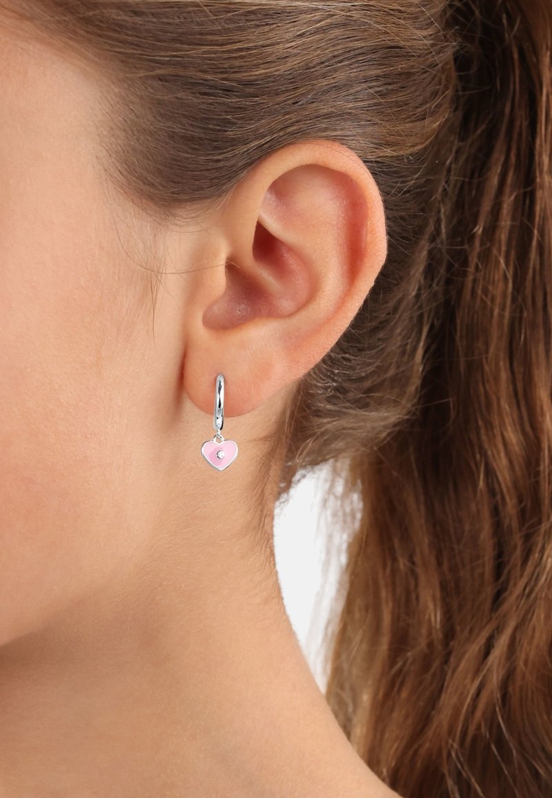 Orecchini a cerchio in argento con un piccolo charm a forma di cuore rosa con un gemma centrale, appesi a un lobo dell'orecchio su uno sfondo neutro.