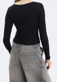 Schwarzes, geripptes langärmliges Oberteil mit einem gewellten Ausschnitt, kombiniert mit locker sitzenden grauen Jeans mit Gesäßtaschen und einer leicht verbleichten Textur.