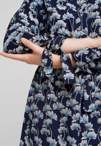 En person som bär en marinblå blommig klänning med vita och ljusblå blommor, med korsade armar och knutna ärmslut.